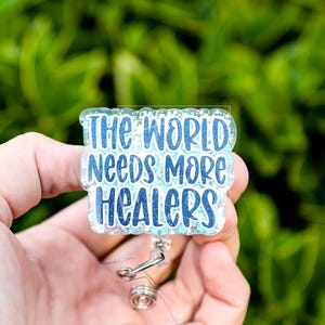 May include: A badge reel with the text "THE WORLD NEEDS MORE HEALERS" in blue lettering. The badge has a glittery, light blue background and a silver retractable clip. The background is a blurred green.
