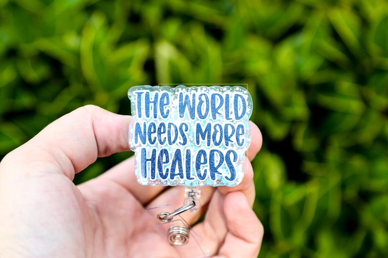 May include: A badge reel with the text "THE WORLD NEEDS MORE HEALERS" in blue lettering. The badge has a glittery, light blue background and a silver retractable clip. The background is a blurred green.