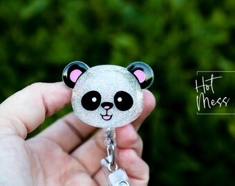 May include: A glittery panda bear badge reel with black and pink accents. The panda has large black eyes and a smiling mouth. The badge reel has a retractable cord and a clip for attaching to clothing. The text "Hot Mess Creative Shop" is in the background.