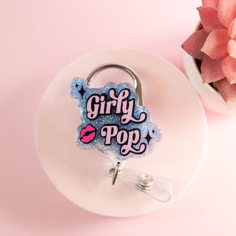 Op de afbeelding: Een glitterende badge reel met de woorden "Girly Pop" in een speels lettertype, met een roze lip-afbeelding. De reel heeft een intrekbaar koord en een metalen clip om aan kleding of accessoires te bevestigen. De achtergrond is zachtroze.
