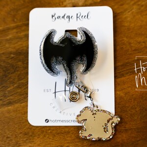 Puede incluir: Un carrete de insignia de murci&eacute;lago negro y plateado con purpurina, con un amuleto de purpurina plateada. El carrete de insignia est&aacute; etiquetado como "Badge Reel" y "Hot Mess Creative Shop".