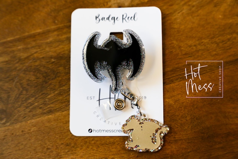 Puede incluir: Un carrete de insignia de murci&eacute;lago negro y plateado con purpurina, con un amuleto de purpurina plateada. El carrete de insignia est&aacute; etiquetado como "Badge Reel" y "Hot Mess Creative Shop".