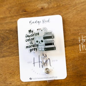 May include: A badge reel with a clear plastic charm featuring a cartoon cat sitting on a stack of books. The charm has the text "My favorite color is morally grey". The badge reel is silver and has a black and white "Hin" logo.