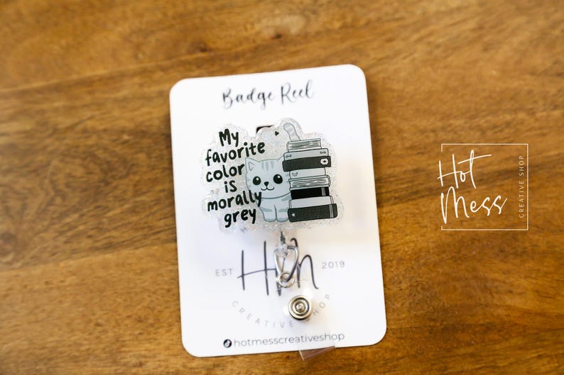 May include: A badge reel with a clear plastic charm featuring a cartoon cat sitting on a stack of books. The charm has the text "My favorite color is morally grey". The badge reel is silver and has a black and white "Hin" logo.