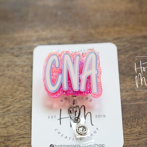 K&ouml;nnte beinhalten: Rosa glitzernder CNA-Badge-Rollenhalter mit silbernem Clip und wei&szlig;em Hintergrund. Der Hintergrund tr&auml;gt den Text "Hot Mess Creative Shop" und "Est 2019".