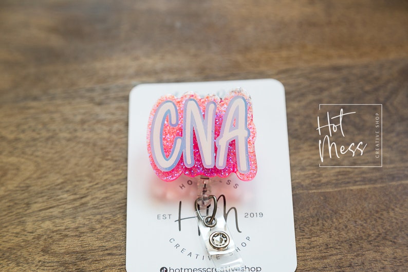 K&ouml;nnte beinhalten: Rosa glitzernder CNA-Badge-Rollenhalter mit silbernem Clip und wei&szlig;em Hintergrund. Der Hintergrund tr&auml;gt den Text "Hot Mess Creative Shop" und "Est 2019".