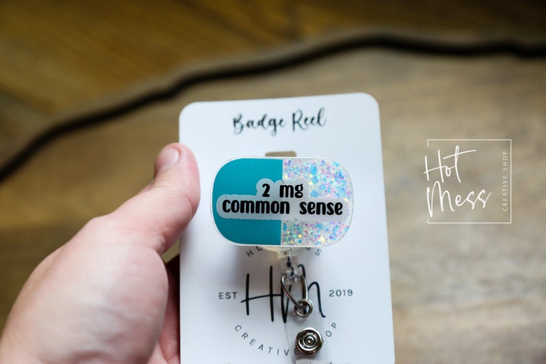 May include: A white card with the text "Badge Reel" and "Hot Mess Creative Shop" printed on it. A teal and white glitter badge reel with the text "2 mg common sense" is attached to the card.
