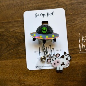Puede incluir: Un portatarjetas con un dise&ntilde;o de OVNI hologr&aacute;fico, un extraterrestre verde con purpurina y un dije de vaca. El portatarjetas est&aacute; sujeto a una tarjeta blanca con el texto "Badge Reel" y "Hot Mess Creative Shop."