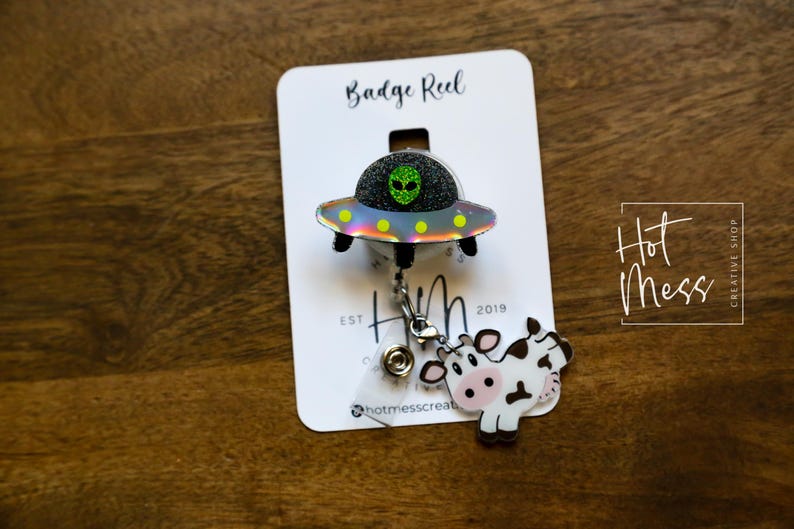 Puede incluir: Un portatarjetas con un dise&ntilde;o de OVNI hologr&aacute;fico, un extraterrestre verde con purpurina y un dije de vaca. El portatarjetas est&aacute; sujeto a una tarjeta blanca con el texto "Badge Reel" y "Hot Mess Creative Shop."
