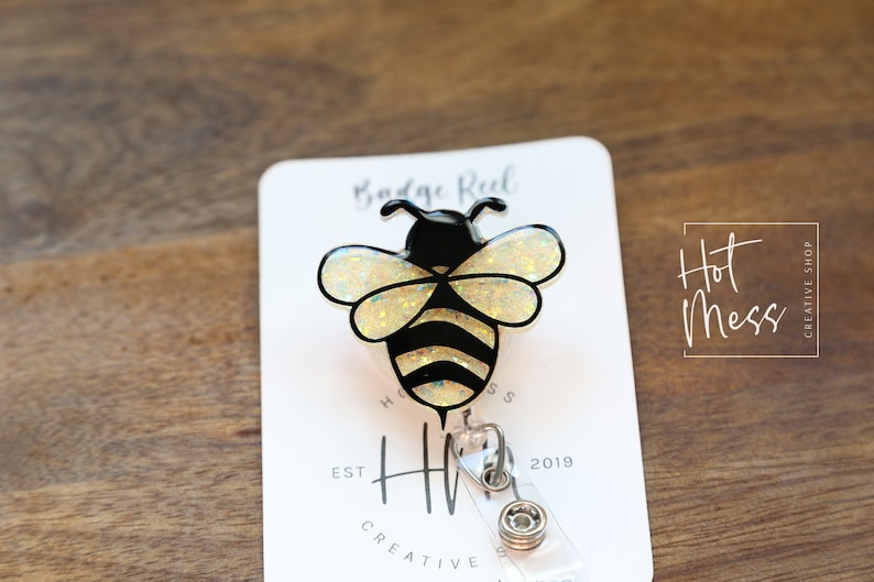 May include: A bee-shaped badge reel with a black and yellow design. The bee has glittery wings and black stripes on its body. The badge reel is attached to a white card with the text "Badge Reel" and "Hot Mess Creative Shop".