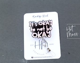 May include: A black and pink badge reel with the text "It's okay to not be okay". The reel is on a white card with the text "Badge Reel" and "Hot Mess Creative Shop".