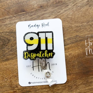 Puede incluir: Un portatarjetas con el texto "911 Dispatcher" en negro, amarillo y blanco. El portatarjetas est&aacute; sujeto a una tarjeta blanca con las palabras "Badge Reel" y "Hot Mess Creative Shop". El fondo es una superficie de madera.