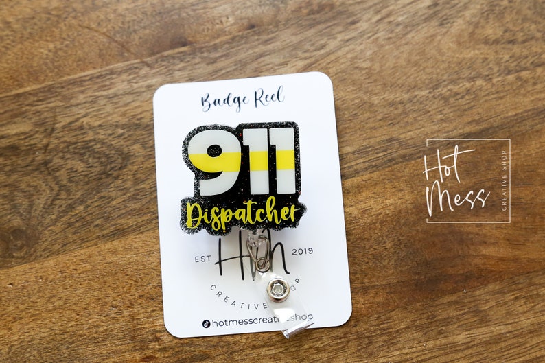 Puede incluir: Un portatarjetas con el texto "911 Dispatcher" en negro, amarillo y blanco. El portatarjetas est&aacute; sujeto a una tarjeta blanca con las palabras "Badge Reel" y "Hot Mess Creative Shop". El fondo es una superficie de madera.