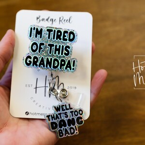 May include: A white card with the text "Badge Reel" and "I'm Tired of This Grandpa!" in black letters with glitter accents. A silver retractable badge reel with a silver charm that says "Well That's Too Dang Bad!" is attached to the card.
