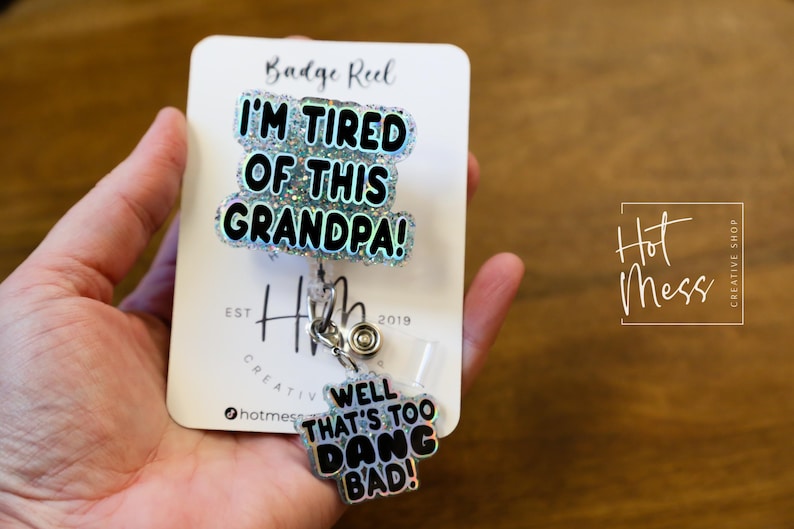 May include: A white card with the text "Badge Reel" and "I'm Tired of This Grandpa!" in black letters with glitter accents. A silver retractable badge reel with a silver charm that says "Well That's Too Dang Bad!" is attached to the card.