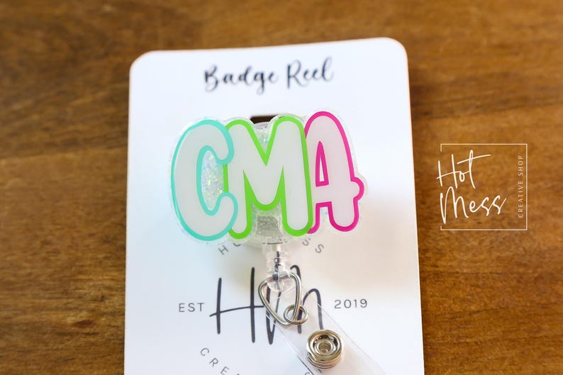 K&ouml;nnte beinhalten: Ein Ausweis-Rollhalter mit den Buchstaben "CMA" in einem Farbverlauf aus Hellblau, Gr&uuml;n und Pink. Der Rollhalter hat einen Stethoskop-Anh&auml;nger und den Text "Hot Mess Creative Shop" und "Est 2019".