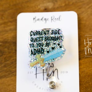 Pode incluir: Um carretel de crachá de glitter prateado com um design de espada azul e branca. O texto no carretel diz "Current Side Quest Brought To You By ADHD".