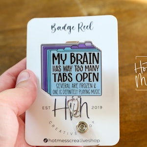 May include: A white badge reel with a colorful design. The design features a stack of file folders with the text "MY BRAIN HAS WAY TOO MANY TABS OPEN". The reel also has the text "SEVERAL ARE FROZEN & ONE IS DEFINITELY PLAYING MUSIC".