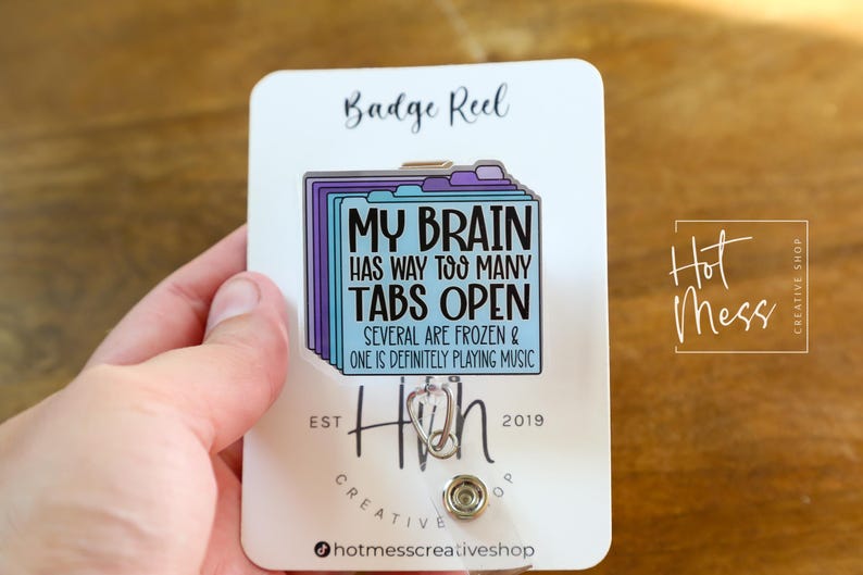 May include: A white badge reel with a colorful design. The design features a stack of file folders with the text "MY BRAIN HAS WAY TOO MANY TABS OPEN". The reel also has the text "SEVERAL ARE FROZEN & ONE IS DEFINITELY PLAYING MUSIC".