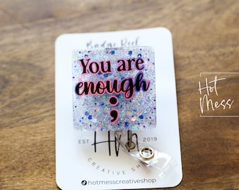 May include: A clear badge reel with a white glitter background and the text "You are enough" in black and pink lettering. A semicolon and comma are below the text. The badge reel is attached to a clear plastic badge holder.