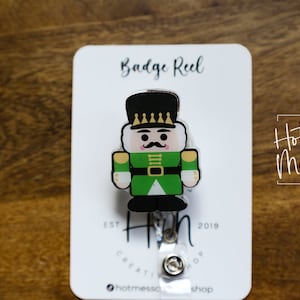 Puede incluir: Un carrete de insignia de cascanueces verde y dorado con fondo blanco. El carrete de insignia tiene el texto "Hot Mess Creative Shop" y "Est. 2019" en él.