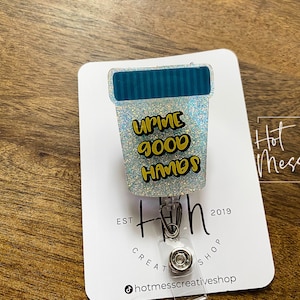 Puede incluir: Un portagafete de purpurina plateada con un diseño de taza azul y blanca. La taza tiene el texto "Upme Good Hands" en letras amarillas. El portagafete está unido a un carrete retráctil de gafete plateado.