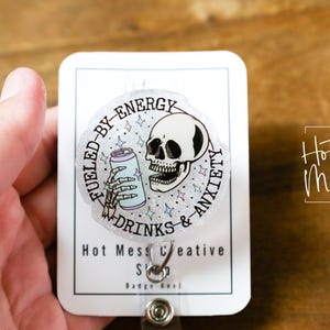 May include: A badge reel with a circular design featuring a skull, a skeleton hand holding a can, and the text "Fueled by Energy, Drinks & Anxiety." The badge reel also has the text "Hot Mess Creative Shop."