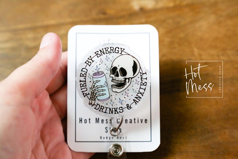 May include: A badge reel with a circular design featuring a skull, a skeleton hand holding a can, and the text "Fueled by Energy, Drinks & Anxiety." The badge reel also has the text "Hot Mess Creative Shop."