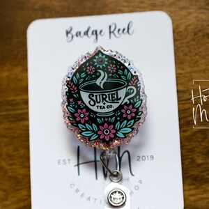 May include: A white and black badge reel with a glitter finish. The design features a cup of tea with the text "Suriel Tea Co." and floral accents. The badge reel is attached to a silver retractable cord.