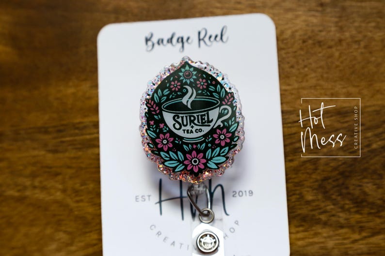 May include: A white and black badge reel with a glitter finish. The design features a cup of tea with the text "Suriel Tea Co." and floral accents. The badge reel is attached to a silver retractable cord.