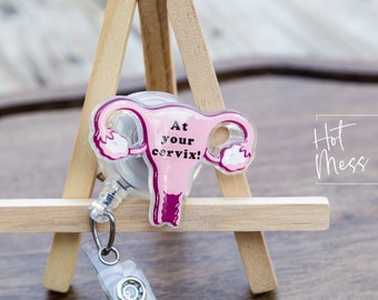 May include: Pink and purple anatomical illustration of a uterus with the text "At your cervix!" on a clear retractable badge reel.