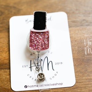 May include: A pink glitter nail polish bottle shaped badge reel with a silver clip and a white card with the text "Hot Mess Creative Shop" and "HM est 2019".