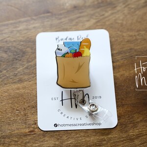 May include: A brown paper bag with a milk carton, apples, bananas, and a loaf of bread inside. The bag is attached to a clear plastic retractable badge holder with a silver clip. The badge holder has a silver charm with the text "Hot Mess Creative Shop" and the text "Est. 2019".