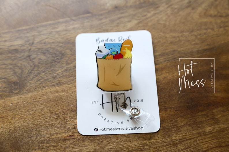 May include: A brown paper bag with a milk carton, apples, bananas, and a loaf of bread inside. The bag is attached to a clear plastic retractable badge holder with a silver clip. The badge holder has a silver charm with the text "Hot Mess Creative Shop" and the text "Est. 2019".