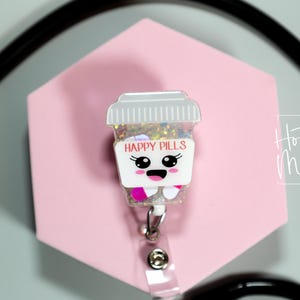 May include: A badge reel with a cartoon pill bottle design, displaying the words "HAPPY PILLS". The badge has a smiling face and is filled with glitter. It is set against a pink hexagonal background.
