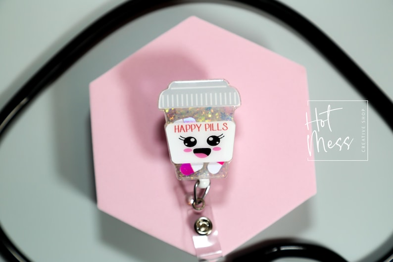 May include: A badge reel with a cartoon pill bottle design, displaying the words "HAPPY PILLS". The badge has a smiling face and is filled with glitter. It is set against a pink hexagonal background.