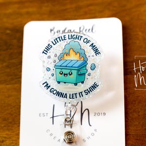 May include: A white glitter badge reel with a cartoon image of a green trash can with a smiling face and flames coming out of it. The text around the image reads "This little light of mine I'm gonna let it shine".