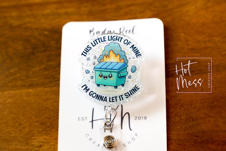 May include: A white glitter badge reel with a cartoon image of a green trash can with a smiling face and flames coming out of it. The text around the image reads "This little light of mine I'm gonna let it shine".
