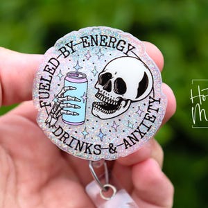 May include: A round, glittery badge reel with the text "Fueled by Energy, Drinks & Anxiety." The design features a skeleton hand holding a light blue can and a skull. The badge reel has a silver clip.