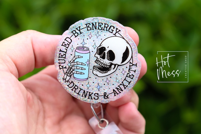 May include: A round, glittery badge reel with the text "Fueled by Energy, Drinks & Anxiety." The design features a skeleton hand holding a light blue can and a skull. The badge reel has a silver clip.