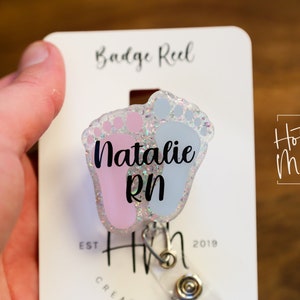 May include: A pink and blue glitter baby footprint badge reel with the name "Natalie RN" printed on it. The badge reel is attached to a white card with the text "Badge Reel" and "Hot Mess Creative Shop" printed on it.
