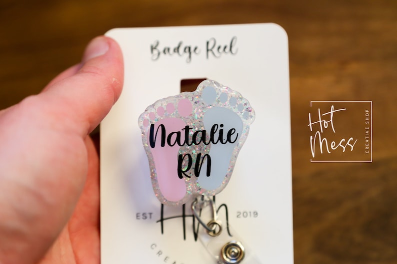 May include: A pink and blue glitter baby footprint badge reel with the name "Natalie RN" printed on it. The badge reel is attached to a white card with the text "Badge Reel" and "Hot Mess Creative Shop" printed on it.