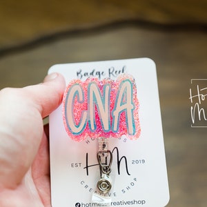 K&ouml;nnte beinhalten: Rosa glitzernder Badge-Halter mit dem Text "CNA" in wei&szlig;en Buchstaben. Der Badge-Halter ist an einer wei&szlig;en Karte mit dem Text "Hot Mess Creative Shop" und "Est. 2019" befestigt.