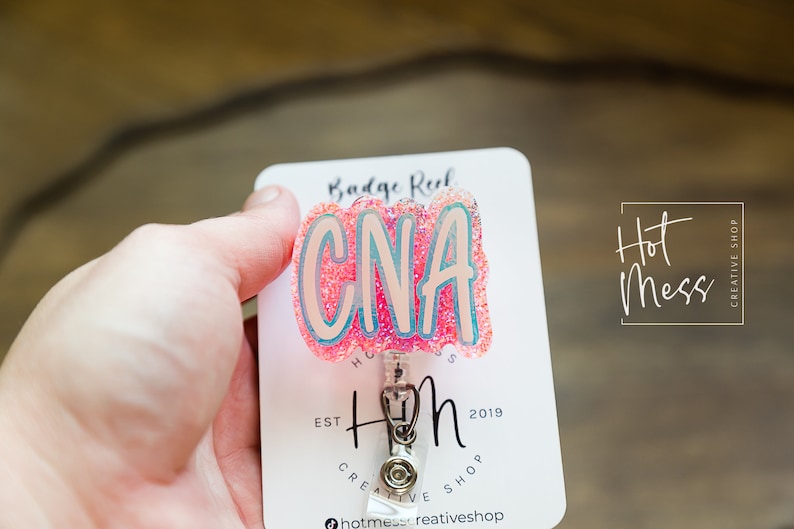 K&ouml;nnte beinhalten: Rosa glitzernder Badge-Halter mit dem Text "CNA" in wei&szlig;en Buchstaben. Der Badge-Halter ist an einer wei&szlig;en Karte mit dem Text "Hot Mess Creative Shop" und "Est. 2019" befestigt.
