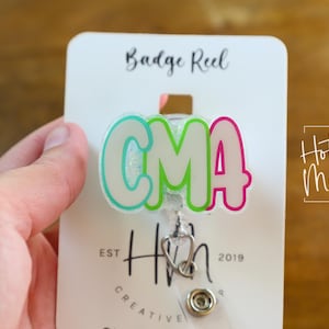K&ouml;nnte beinhalten: Ein Ausweis-Rollhalter mit den Buchstaben "CMA" in einer Blasen-Schriftart, umrandet in T&uuml;rkis und Pink, mit einem Stethoskop-Anh&auml;nger. Der Ausweis-Rollhalter ist an einer wei&szlig;en Karte mit den Worten "Badge Reel" und "Hot Mess Creative Shop" befestigt.