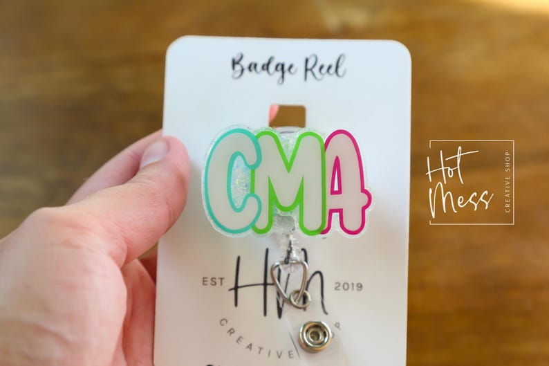 K&ouml;nnte beinhalten: Ein Ausweis-Rollhalter mit den Buchstaben "CMA" in einer Blasen-Schriftart, umrandet in T&uuml;rkis und Pink, mit einem Stethoskop-Anh&auml;nger. Der Ausweis-Rollhalter ist an einer wei&szlig;en Karte mit den Worten "Badge Reel" und "Hot Mess Creative Shop" befestigt.