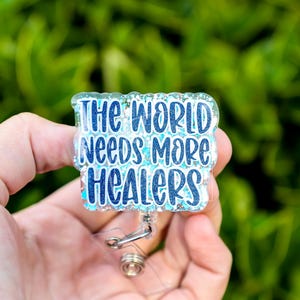 May include: A rectangular badge reel with the text "THE WORLD NEEDS MORE HEALERS" in blue and white lettering. The badge has a glittery, clear border and a retractable cord with a metal clip.