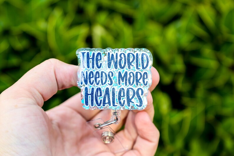 May include: A rectangular badge reel with the text "THE WORLD NEEDS MORE HEALERS" in blue and white lettering. The badge has a glittery, clear border and a retractable cord with a metal clip.