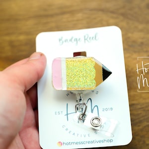 May include: A gold glitter pencil badge reel with a pink eraser. The badge reel is attached to a clear plastic retractable badge holder. The badge reel is on a white card with the text "Badge Reel" and "Hot Mess Creative Shop" printed on it.