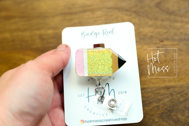 May include: A gold glitter pencil badge reel with a pink eraser. The badge reel is attached to a clear plastic retractable badge holder. The badge reel is on a white card with the text "Badge Reel" and "Hot Mess Creative Shop" printed on it.